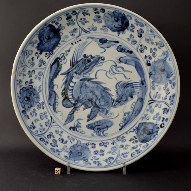 A Ming Blue and White Porcelain dish, Hongzhi 1488 – 1505 or Zhengde 1506 – 1521 period, Jingdezhen kilns. Painted in a bold style using a wet brush with a large Qilin in a landscape with a border of flowering peony.Condition Poor ; four large rim cracks, the surface is a bit worn through general use. The tone of blue is slightly paler than in the photograph. Size Diameter : 12 inches (31 cm) Provenance Giangarlo Folco Collection (label to the base) Stock number 24617