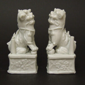 A Pair of Kangxi Blanc de Chine Porcelain `Dogs of Fu` Taper-Stick Holders. The Sides Decorated with Flowering Prunus in Relief. From a Kiln in Dehua, Fujian Province, Kangxi Period 1662 - 1722. Robert McPherson Antiques - Sold Archive 22987.