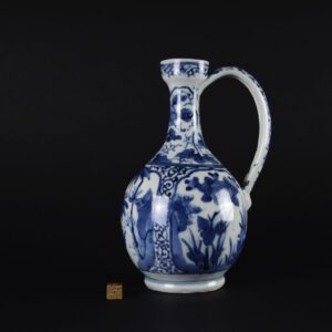 A 17th Century Japanese Export Porcelain Jug or Ewer, Arita Kilns c.1670-1690. This elegant Japanese blue and white porcelain ewer is somewhat of a hybrid. The shape is Dutch in origin, and 17th century Dutch Delft examples of this ewer are well known, see 'References'. The painting style is a mixture of Japanese, with influences of contemporary Chinese Transitional porcelain as well as Dutch Delftware. The bulbous lower section of the jug is divided into three panels, these are framed with Karakusa scrolls. One panel lined up below the spout is of a couple wondering around in a rather fantastical landscape, the lady has her parasol open while the man’s eye has been caught by something of interest. Large bamboo leaves and curious rocks fill the scene. The panels that flank this scene are of flowering peony and other flowers. The slender high handle has a hole at the top to affix a European mount and cover. The tapering neck flares at the top, which is slightly pinched to form a very minimal, rounded spout. Two sizes of this export porcelain jug are known, this being the smaller of the two. The larger size is around 28cm. SOLD