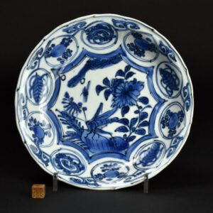 A Late Ming Kraakware Saucer Shaped Dish c.1610 – 1630. The center of the dish, decorated in tones of cobalt, decorated with a grasshopper on a rock next to a flowering peony. The cavetto of this thickly potted Kraak porcelain dish has ten peach shaped panels filled with flowers, peaches and auspicious objects. The base has feint chatter marks and grit adhering to the glaze. The grit was used to stop the piece sticking in the glaze firing.For more information about Kraak Ware porcelain you can go to the History section of our website-menu, or click on the link: Kraak Ware Porcelain SOLD Condition A very small glaze chip to the back, very minor friting, minor wear,a few small pieces of kiln grit stuck to the well of the dish. Size Diameter 20 cm (8 inches) Provenance An old American dealers label (discoloured) with a stock number and price. Stock number 25602