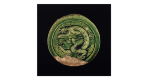 Museum number1933,0317.3 Description Earthenware 'goutou' tile terminal with moulded decoration beneath a green glaze. This circular 'goutou' tile terminal has a raised border 2 cm wide and a relief-moulded dragon in the centre, covered with a bottle-green glaze. The dragon has a scaly sinewy body and paws with five flexed claws. It twists round, prancing on its front legs and turning its back legs under and over its tail. It is damaged along the lower edge and is missing its convex section.