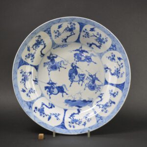 A Large Kangxi Blue and White Porcelain dish, the base with an apocryphal Chenghua mark (the Ming Emperor Chenghua reigned from 1465 to 1487). The central scene is of a dynamic Chinese cavalry, four mounted soldiers in armour with plumed helmets are surrounding another mounted soldier who has a smaller plum on his helmet. The border is of flowering prunus in panels reminiscent of Kraakware. Branches of plum blossom convey the `Five Blessings` Meikai Wufu – longevity, wealth, health, love of virtue and a peaceful death.Condition In good condition but with some small rim chips. There is an area of deep fritting to the rim c.2cm. Two small shallow (mostly glaze glaze) chips to the front and a minute frit. The back rim has several small chips and glaze frits. There is some very feint speckling to the glaze from the torso of the warrior at 11 o’clock to the rim. Size Diameter 34.5 cm (13 1/2 inches) Provenance From a Private Belgium Collection Stock number 25848