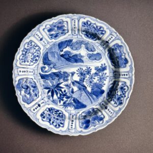 Porcelain dish with 'kraak'-type underglaze blue decoration. This finely potted dish has shallow moulded lobed sides, a flattened rim with lobed edge and a low tapering foot with some grit attached. The central medallion depicts a pheasant perched on a rock beside water and stylized flowering plants and foliage. Surrounding this are eight panels containing cartouches with opposite pairs of chrysanthemum, morning glory, butterfly, possibly camellia, bordered by narrow panels with dots and lozenges or dots and spirals. The reverse is painted with alternating panels with butterflies or peaches. The glazed base is marked with a standing egret in underglaze blue in the centre.
