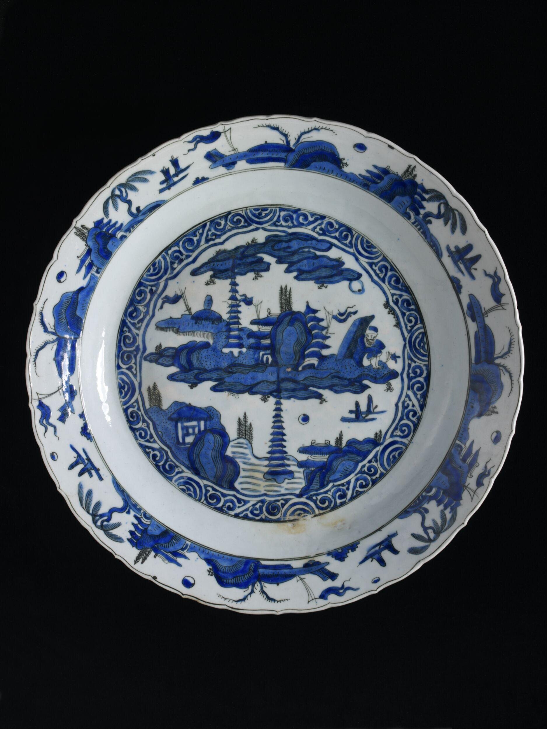Dish of fritware, painted in two shades of blue with black outline under a clear glaze. In the central medallion is a landscape with pagodas, islands and boats, framed by scrollwork. A border with repeated landscape design is depicted round the flattened and foliated rim. There is an imitation Chinese square mark in black.