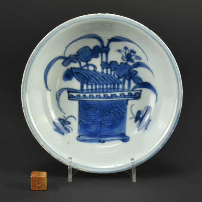 A Late Ming Porcelain Bowl, Later Wanli or Tianqi c.1600 – 1627, Jingdezhen Kilns. This small Ko-Sometsuke (old blue and white) bowl with a thick base and thin everted rom is decorated with flowering lotus from a plant pot, either side is a small display of a scholars rock in a pot with grasses. The bases has been cut out, leaving chatter-marks where the finishing tool has juddered, rather than finishing of the base neatly. The base has been left unglazed, the footrim has grit stuck to it, it would have been fired while sitting on this grit. This rough, uneven accidental texture contrasts with the silky surface of the porcelain, something that would have great appeal to the Japanese. Read more about Ko-Sometsuke porcelain below.See Below For More Photographs and Information. Condition Mushikui (insect-nibbled) small rim chips, typical of this Ming porcelain for Japan. Size Diameter 14 cm (5 1/2 inches) Provenance N/A Stock number 26194 £ 340 