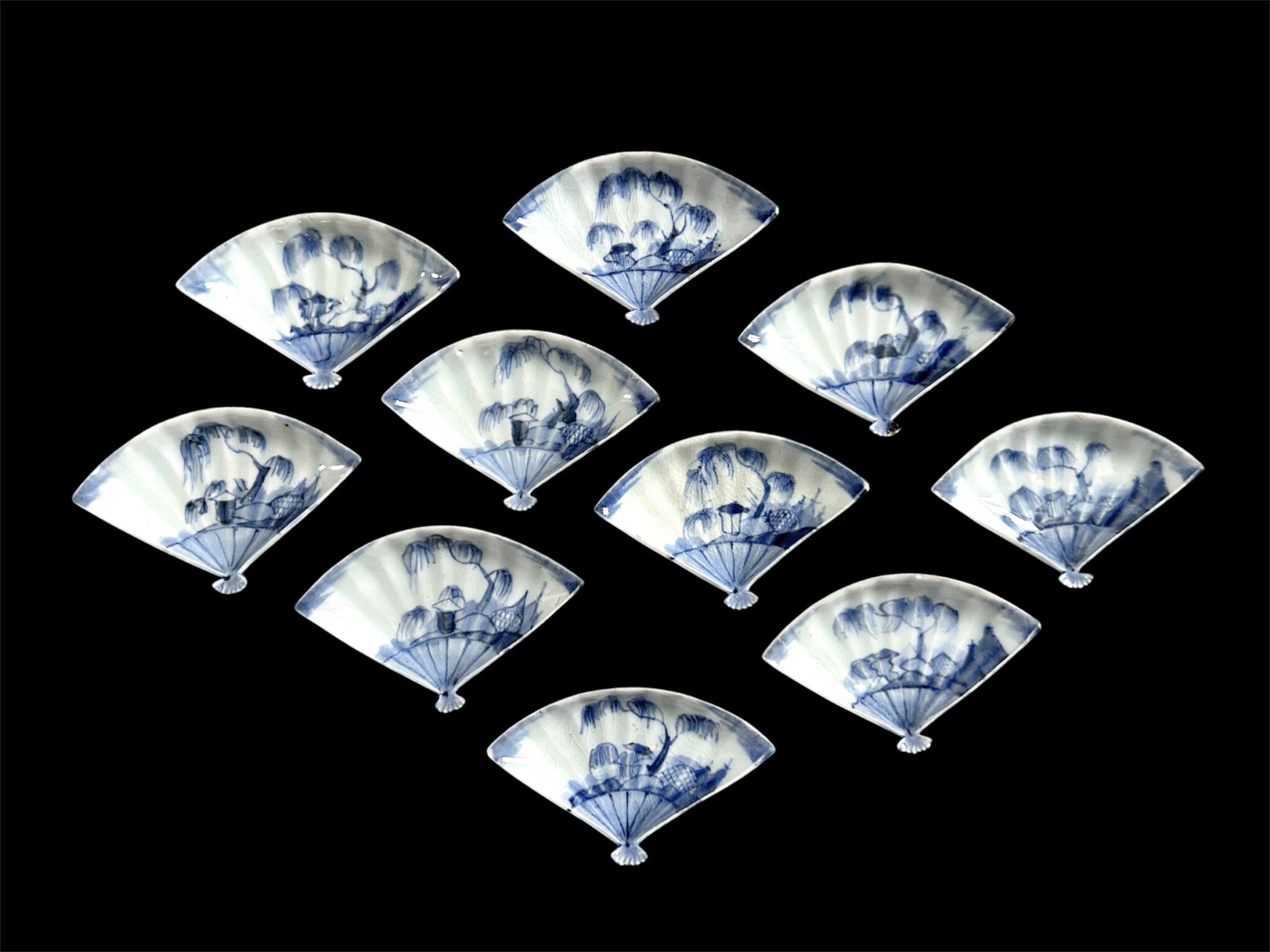 A Rare Set of Ten Small Japanese Blue and White Fan Shape Dishes, Arita Kilns c.1690 – 1720. These very small thinly potted Japanese porcelain dishes are mould made to represent an open fan. The scene fits onto what would be the paper part on the fan. It shows a water side with a large willow tree and a small building, a rustic shelter of some kind, next to that is a bound rock and fishing nets hung to dry. The reverse has an imitation four character Ming mark in cursive script. The H type design represents good luck, its very thin, perhaps suggesting pine needles. It’s rare to find a complete set of ten Japanese porcelain dishes complete with their box. The dishes are in an extensively inscribed Tomobako box.SOLD See Below For More Photographs and Information Condition One dish has two tiny chips, Six of these tiny Japanese porcelain dishes have one tiny chip, Only three are perfect. Size Diameter 9.9 cm (3.9 inches), depth 2 cm (0.8 inches). Provenance N/A Stock number 27551 References For a single porcelain dish of this size, shape and design see : Complete Catalogue of Shibata Collection (Contributors Saga Kenritsu Kyūshū Tōji Bunkakan. Published by the Kyushu Ceramic Museum, 2019) page 314. Item 2502. The Shibata dish has a difference, the back design is of pine needles.