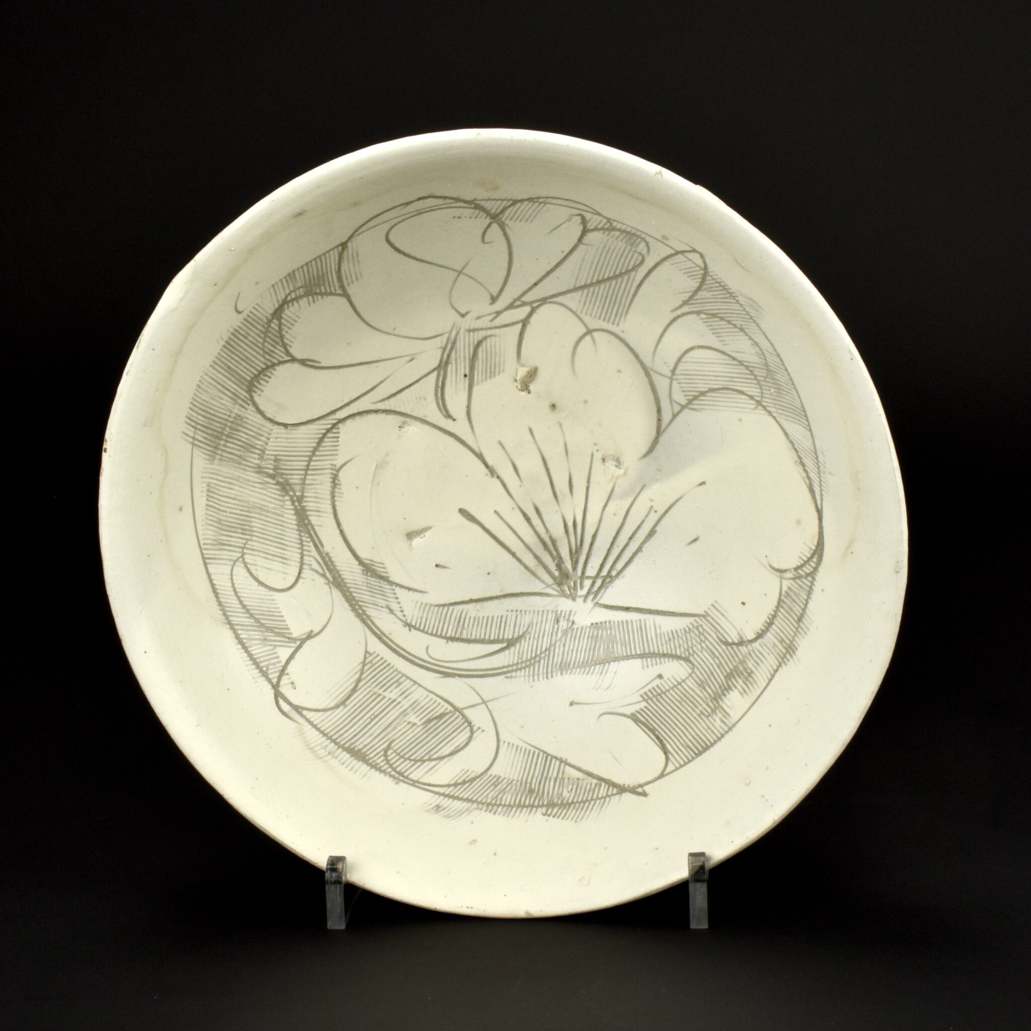 A Cizhou Type Carved and Combed Stoneware Bowl, Northern Song c.12th Century, probably from the Guantai kilns in Handanxian or a kiln Hebiji, Tanqyinxian. The stoneware bowl with sgraffito decoration of open peony flowers that overshoot the single incised border line. The putty-grey coloured stoneware body has a creamy white slip applied that has been fluidly incised using a stylus and a comb. The piece was then given an uneven thin clear glaze, the dipped slip is also somewhat uneven in depth. There are spur-marks visible to the well of the bowl and as grey shadows on the footrim from the kiln furniture. These bowls were piled up, they must have had a group sagger, rather than individual one.